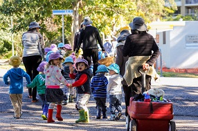 Children and educators walking down a leafy street while pulling a red wheelbarrow