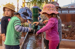 Four children wearing hats playing outdoors with an old-fashioned water pump and tray