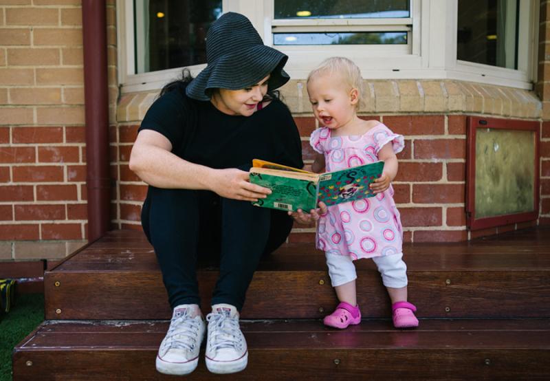 Educator sitting on steps and reading book with toddler